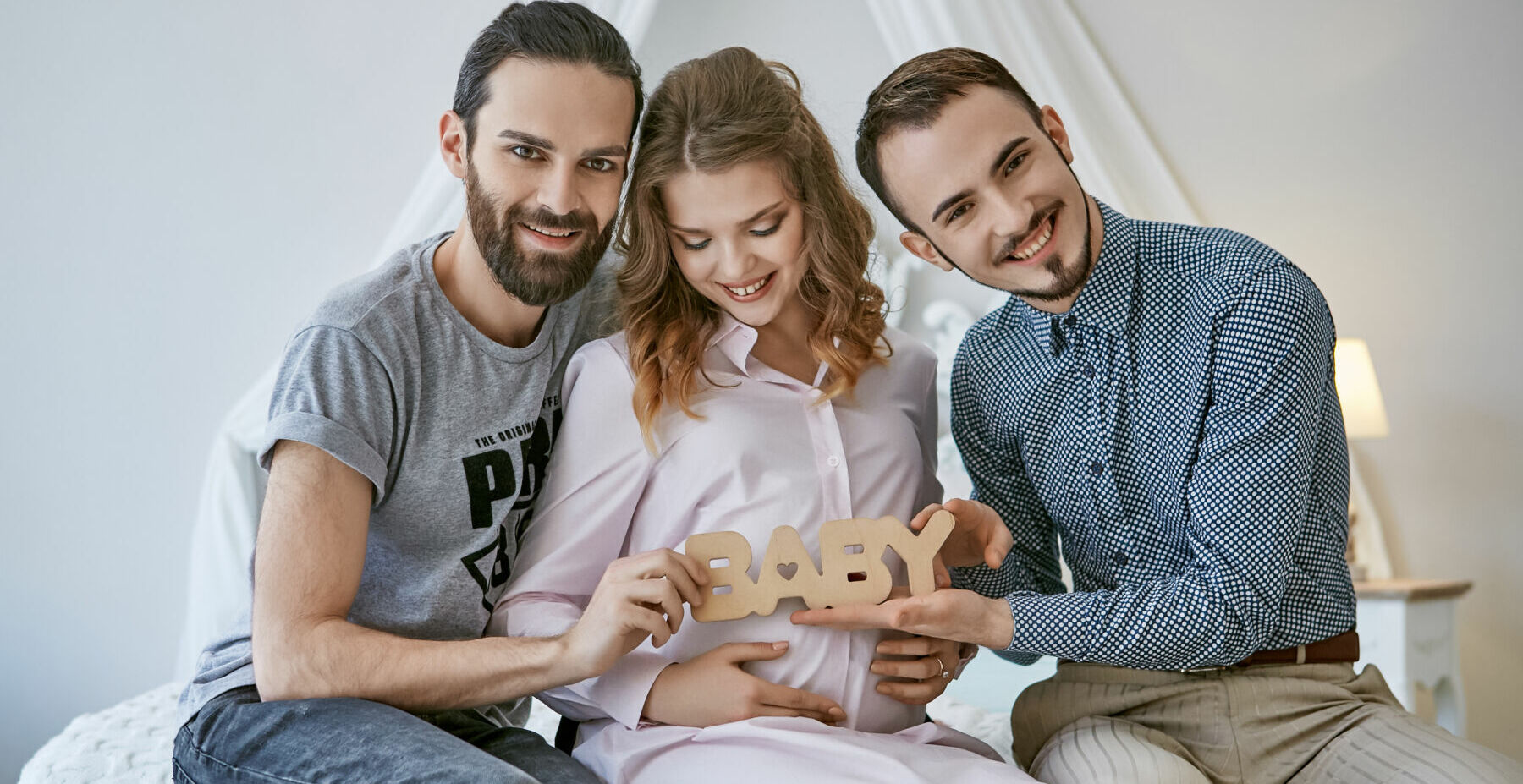 Gay couple becoming parents through surrogacy. The fathers-to-be sitting on the bed, a surrogate mom between them, happy smiles on their faces. The men holding lettering BABY over the pregnant belly.
